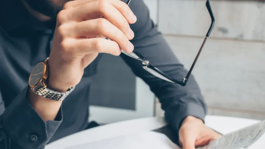 Man holding a pair of glasses wearing a watch and holding a paper document