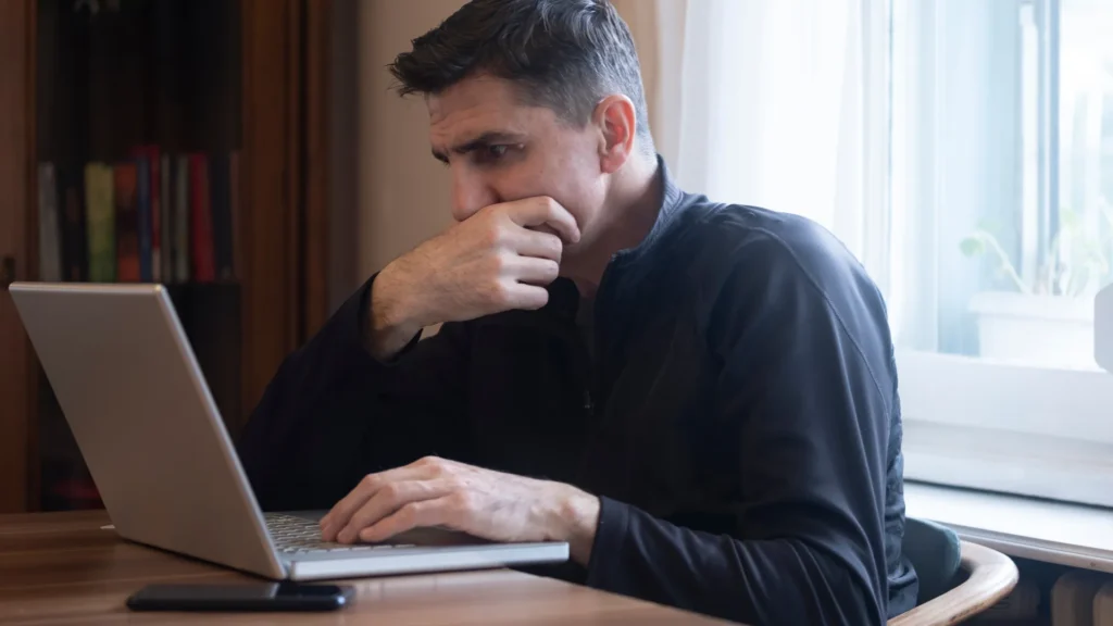 A stressed man wearing black sitting at a table with his laptop open