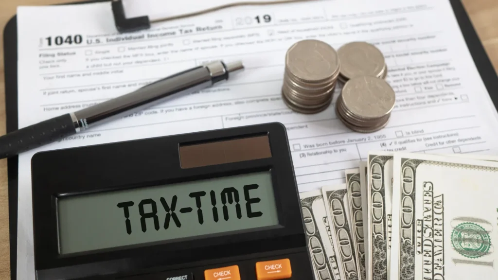 Calculator that says 'tax-time' sitting on top of tax documents with cash, coins, and a pen next to it