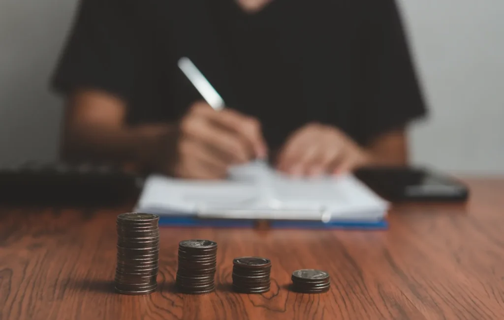 4 stacks of coins, each one smaller in size in foreground with man i background with pen in his hand