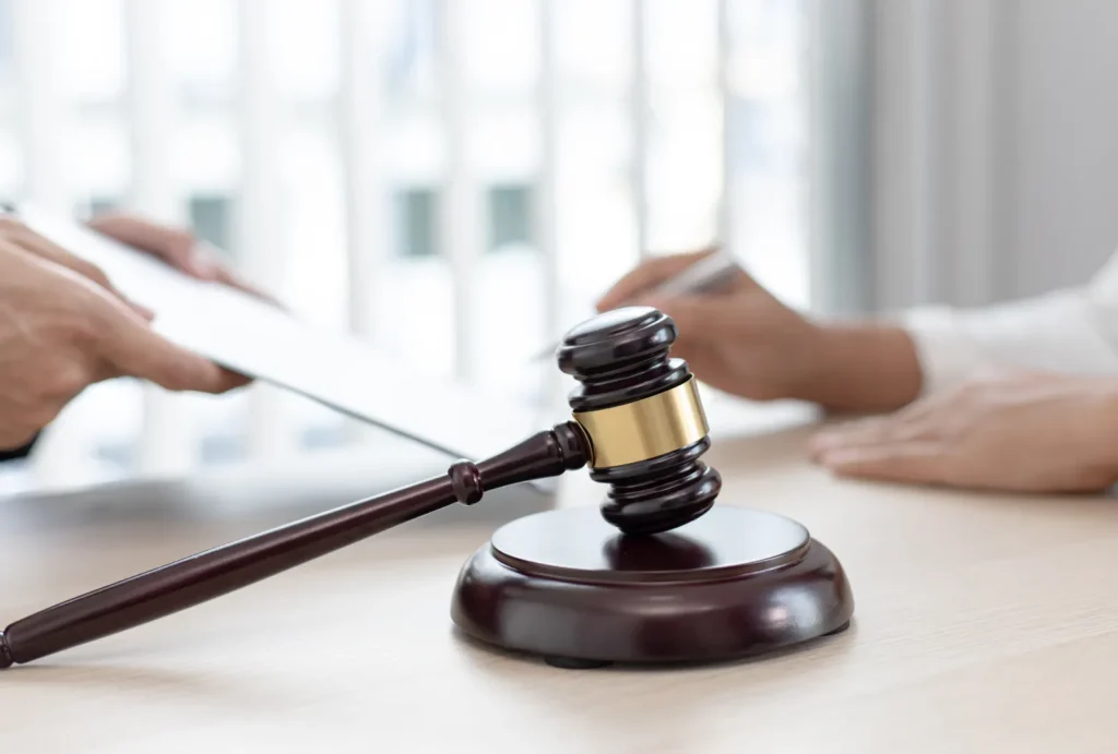 attorney gavel sitting on a desk in the foreground with one person signing a document in the background